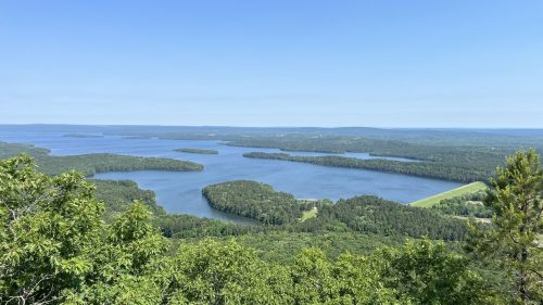A panoramic view of a serene lake surrounded by lush greenery and distant hills under a clear blue sky.