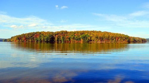 A serene lake scene with calm waters reflecting an island surrounded by colorful autumn foliage under a clear blue sky.