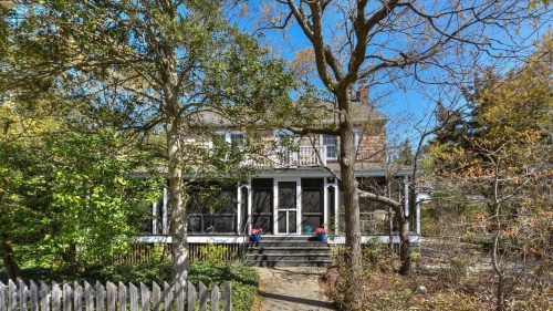A charming two-story house surrounded by trees and a picket fence, with a porch and colorful flower pots.