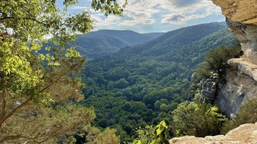 A scenic view of lush green mountains and valleys framed by rocky cliffs under a bright sky.