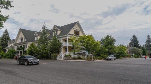 A large, multi-story house with a steep roof, surrounded by trees, at a street intersection on a cloudy day.
