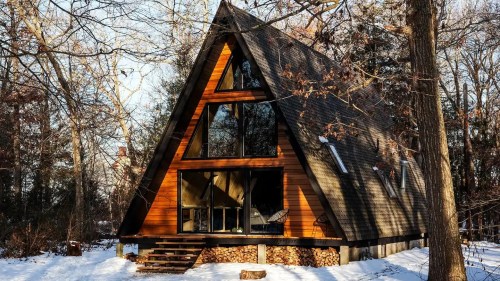 A cozy A-frame cabin surrounded by snow-covered trees, featuring large windows and a woodpile stacked beneath.