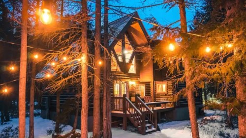 A cozy cabin surrounded by trees, illuminated by string lights, with snow on the ground and a twilight sky.
