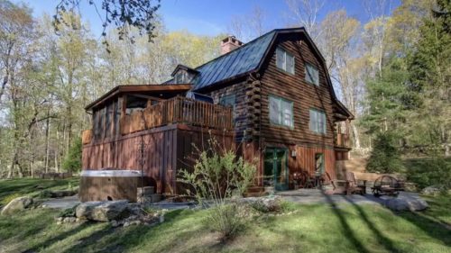 A rustic wooden cabin surrounded by trees, featuring a deck and hot tub in a serene outdoor setting.