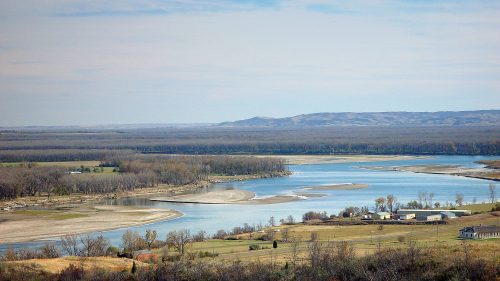 A serene river winding through a landscape of trees and fields under a blue sky with scattered clouds.