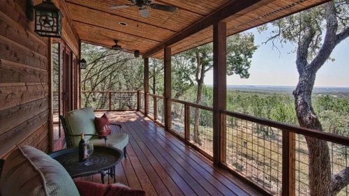 A wooden porch with comfortable seating, overlooking a scenic view of trees and hills under a clear blue sky.