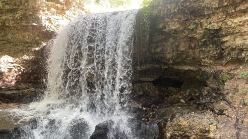 A cascading waterfall flows over rocky terrain, surrounded by lush greenery and natural stone formations.