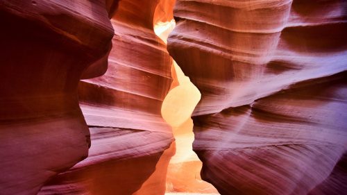 A narrow slot canyon with smooth, colorful rock walls and light streaming through the opening.