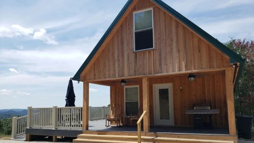 A wooden cabin with a porch, grill, and deck, set against a scenic landscape under a blue sky.