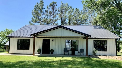 A modern single-story house with a metal roof, surrounded by green grass and trees, featuring a front porch with chairs.