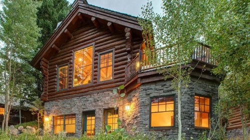 A cozy log cabin with large windows, surrounded by greenery and trees, showcasing warm interior lighting.