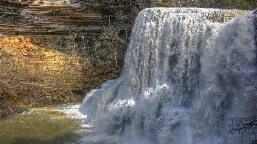 A cascading waterfall flows over rocky cliffs into a serene pool below, surrounded by trees and natural scenery.