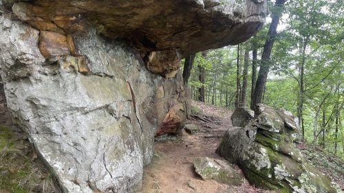 A rocky outcrop with a large overhanging boulder, surrounded by trees and a dirt path in a forested area.