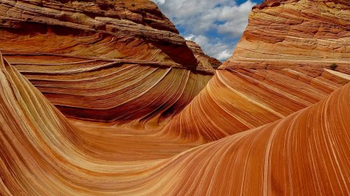 Colorful, wavy rock formations under a blue sky with clouds, showcasing vibrant orange and red stripes.