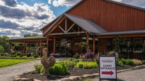 A rustic red barn with a sign pointing to the event entrance, surrounded by colorful flowers and greenery.