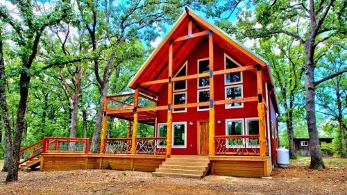 A red cabin with a steep roof, surrounded by trees, featuring a wooden deck and steps leading to the entrance.