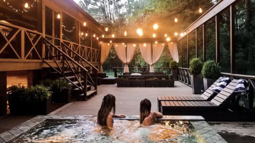 Two people relax in a hot tub on a deck, surrounded by string lights and a serene outdoor setting.