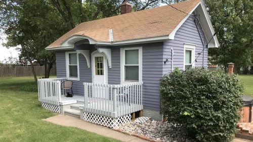 A small purple house with a front porch, surrounded by grass and shrubs, on a sunny day.