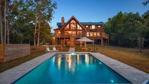 A spacious wooden house with large windows, surrounded by trees, featuring a pool in the foreground at dusk.