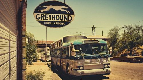 A vintage Greyhound bus parked under a sign in Lowell, Arizona, with a sunny, desolate street scene.