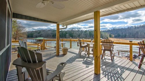 A spacious deck with wooden chairs overlooks a serene lake and wooded landscape under a partly cloudy sky.