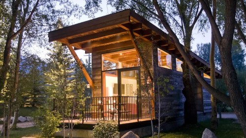 A modern wooden cabin surrounded by trees, illuminated at dusk, with a balcony and large windows.