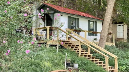A small white cottage with a red roof, surrounded by greenery and pink flowers, featuring a wooden staircase and porch.