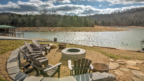 A serene lakeside view with a fire pit and wooden chairs, surrounded by trees and a cloudy sky.