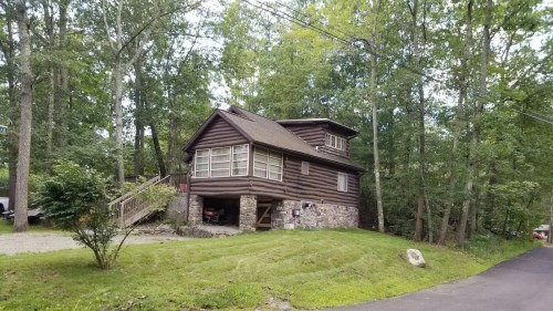 A rustic log cabin surrounded by trees, with a stone foundation and a winding driveway.