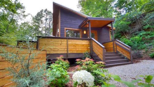 A modern wooden cabin surrounded by greenery, featuring a deck and steps leading to the entrance.