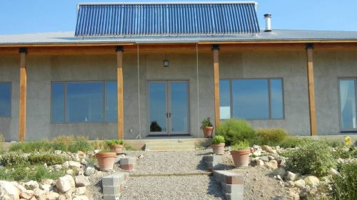 A modern house with a metal roof, large windows, and a stone pathway surrounded by plants and gravel.