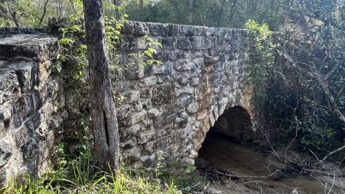 A stone bridge with an arch over a small stream, surrounded by trees and greenery.