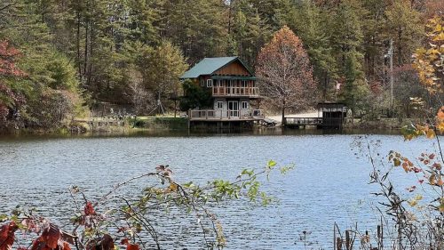 A serene lake scene featuring a wooden house surrounded by trees with autumn foliage. Clear blue sky above.