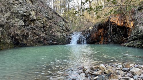A serene, turquoise pool with a small waterfall surrounded by rocky terrain and trees in a peaceful natural setting.