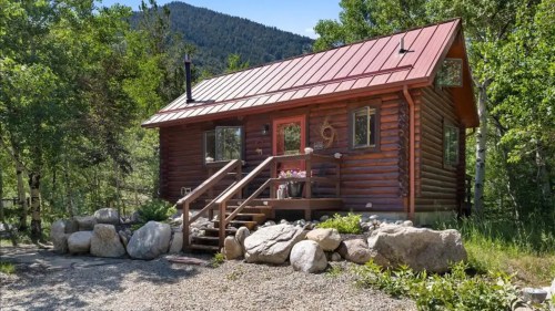 A cozy log cabin with a red metal roof, surrounded by trees and boulders, set against a mountain backdrop.