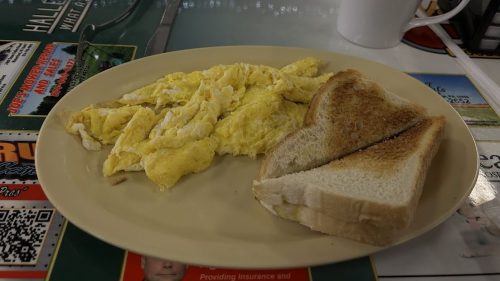 A plate with scrambled eggs and two slices of toasted bread, next to a white coffee mug on a table covered with menus.