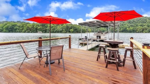 A wooden dock by a lake with red umbrellas over tables and chairs, surrounded by green trees and a blue sky.