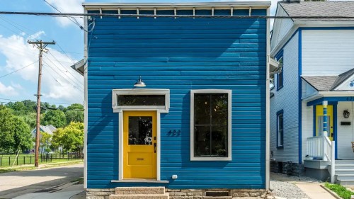 A bright blue house with a yellow door and large window, set against a clear sky and green trees in the background.