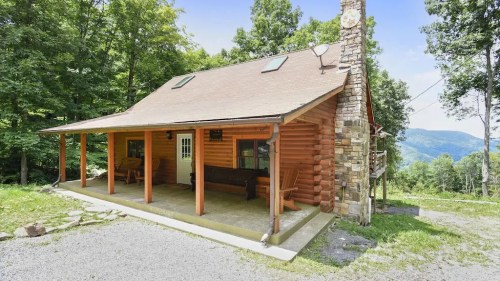A cozy log cabin with a stone chimney, surrounded by trees and mountains, featuring a porch and outdoor seating.