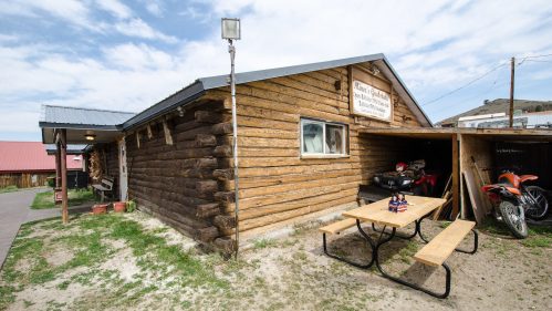 A rustic wooden building with a sign, picnic table, and parked motorcycles in a rural setting under a cloudy sky.
