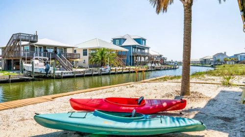 Three kayaks on a sandy shore beside a calm canal, with houses and palm trees in the background under a clear blue sky.