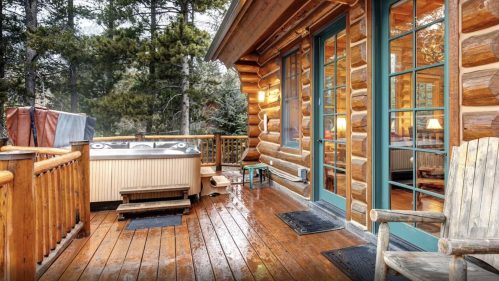 A cozy log cabin deck featuring a hot tub, wooden flooring, and surrounded by trees.