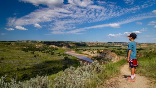 A child stands on a path overlooking a scenic landscape with hills, a river, and a blue sky filled with clouds.