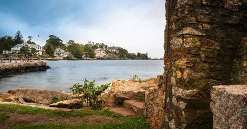 A collection of ruins by the side of a bay, with fancy houses across the water.