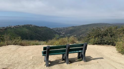 A green bench on a sandy overlook with a view of hills and the ocean under a cloudy sky.