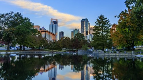 City skyline reflecting in a calm pond, surrounded by trees and colorful autumn foliage under a blue sky.