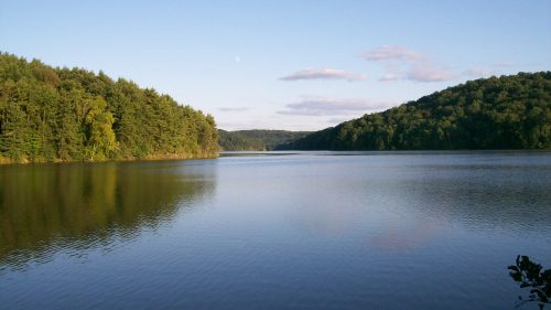 A serene lake surrounded by lush green hills under a clear blue sky with a few clouds.
