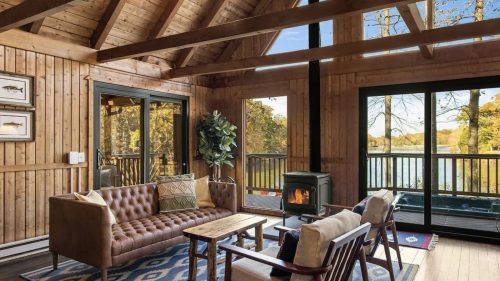 Cozy wooden cabin interior with a sofa, chairs, and a view of a lake through large windows.