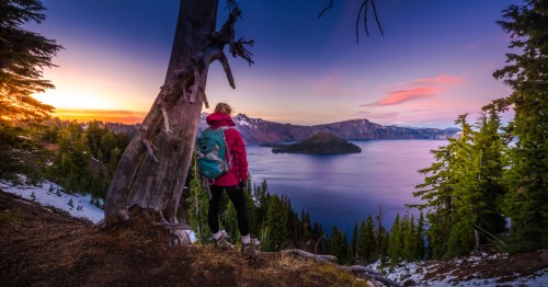 A hiker wearing a pink jacket and a blue backpack stands next to a tree at a secret overlook at Crater Lake National Park in Oregon.