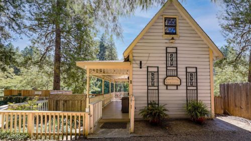 A charming, small white house with a porch, surrounded by tall trees and greenery.